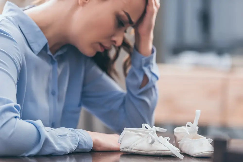 A woman, with her head in her hands, looks down at a pair of baby shoes, expressing a moment of deep thought or concern