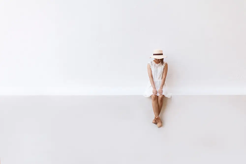 A woman is perched on a wall, legs crossed, appearing thoughtful and at ease in a tranquil outdoor scene.