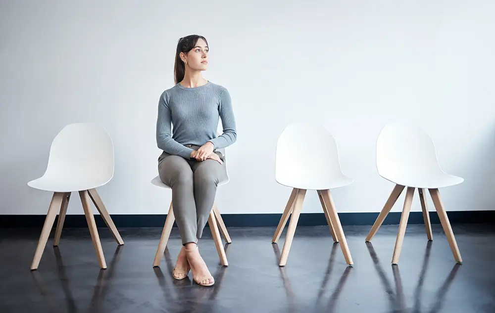 A woman in casual attire sitting on a chair, positioned in front of several empty chairs.