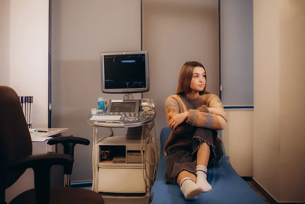 A woman seated on a hospital bed, appearing thoughtful amidst a clinical environment.